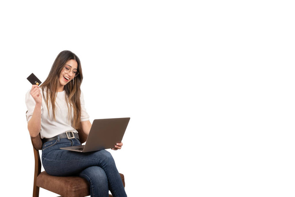 Holding credit card, happy cheerful young brunette caucasian girl holding credit card. Sitting on the chair, using laptop. Internet banking, online shopping concept idea. Surprised, looking computer. - Photo, Image