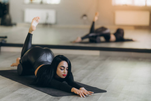 A woman in black sportswear lies on the floor on a mat practicing an asana at home, helping to relieve back pain, a flexible body stretch for beginners. - Fotografie, Obrázek