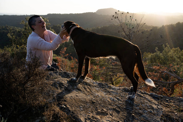 niet-binaire persoon in de zonsondergang met een hond midden in de natuur, bovenop een hondenrots en een vrouw met zeer kort haar met een breed landschap van bossen en velden op de achtergrond. - Foto, afbeelding
