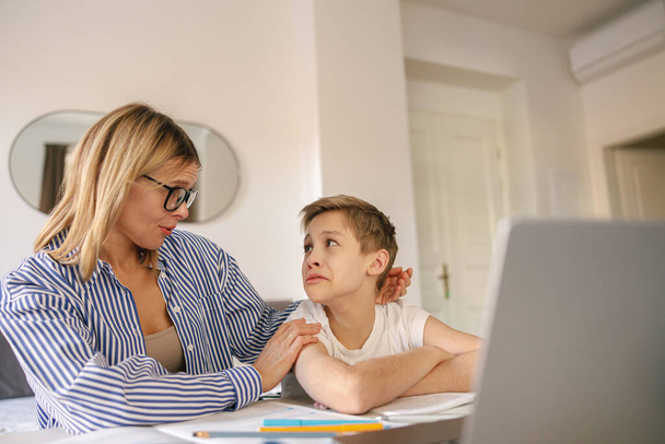 Annesi ona ödevlerinde yardım ederken yorgun, üzgün çocuk dizüstü bilgisayarla masada oturuyor. Yüksek kalite fotoğraf - Fotoğraf, Görsel