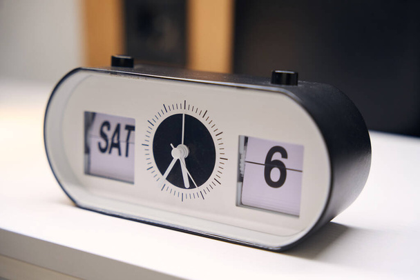 На тумбочці є сучасний електронний будильник, на дисплеї Субота, шостий день
 - Фото, зображення