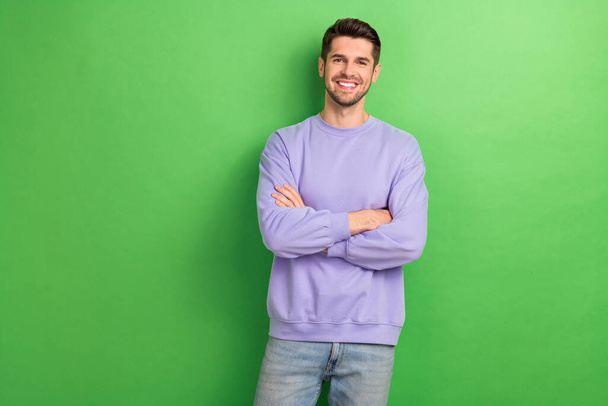 Фото позитивного впевненого молодого чоловіка у фіолетових усміхнених обіймах, схрещених ізольованим зеленим кольором фону
. - Фото, зображення