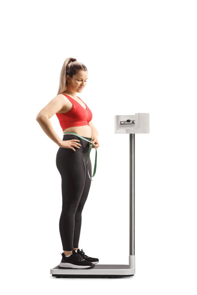 Young woman in sportswear standing on a medical weight scale and measuring her waist isolated on white background - Foto, Imagem