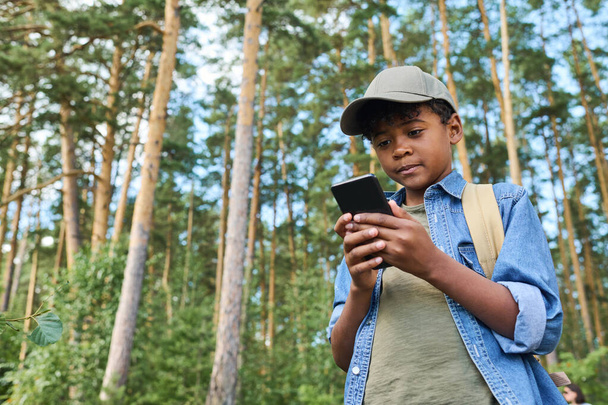 Маленький хлопчик з рюкзаком дивиться карту навігатора в смартфоні, стоячи в сосновому лісі і шукаючи правильний напрямок
 - Фото, зображення