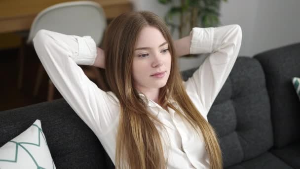 Fiatal szőke nő nyugodt kezekkel a fején ül a kanapén otthon - Felvétel, videó