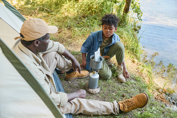 水のそばに座って料理をするのを待っている間に彼の祖父と話す若いアフリカ系アメリカ人の少年 - 写真・画像