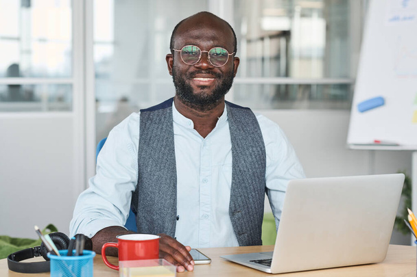 Щасливий молодий афроамериканський менеджер, який працює в мережі перед ноутбуком, сидячи за столом у офісі і дивлячись на камеру - Фото, зображення