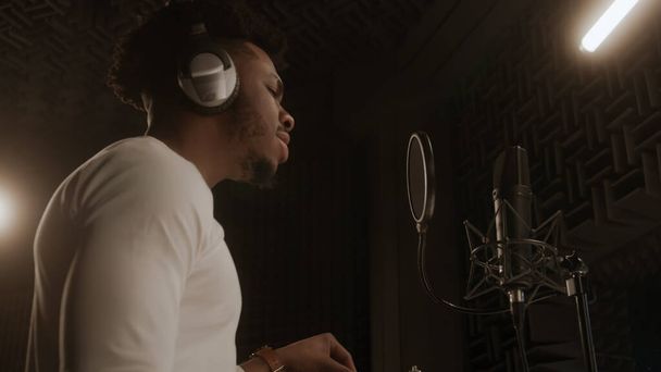 ヘッドフォンのプロの男性ボーカルは、防音室でマイクに叙情的な組成物を歌います。アフリカ系アメリカ人の歌手は録音スタジオで働いている。音楽制作の概念。サイドビュー. - 写真・画像