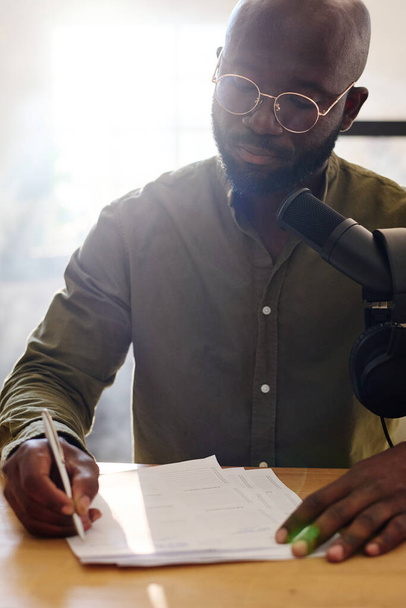 Joven hombre negro calvo leyendo preguntas en papel preparado para la entrevista con el huésped mientras está sentado junto al escritorio en el estudio frente al micrófono - Foto, Imagen