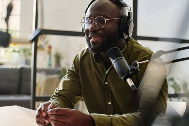 Joven hombre negro en auriculares y anteojos hablando en micrófono mientras está sentado en el lugar de trabajo y la grabación de archivos de audio para su audiencia - Foto, Imagen