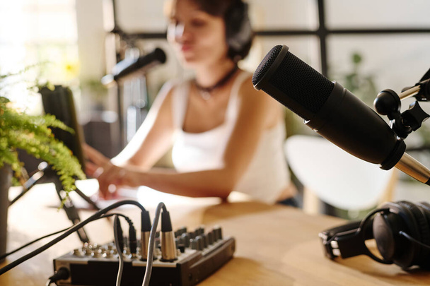 Черный микрофон и другое оборудование для записи аудиофайлов для подкастинга на рабочем месте хоста с девушкой на заднем плане - Фото, изображение