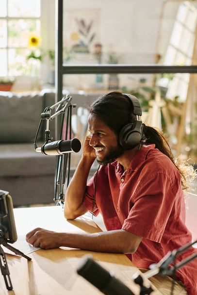 Счастливый молодой человек с темными длинными волосами записывает новые аудио или видео для онлайн аудитории по столу в студии, разговаривая в микрофоне - Фото, изображение