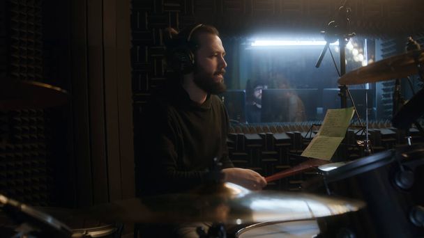 Male musician in headphones plays a drum set. Drummer records song in soundproof music recording room. Audio engineer on background. Work in the sound recording studio. Concept of music production. - Photo, Image
