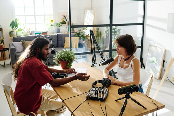 Twee jonge interculturele mensen die discussiëren over populair onderwerp in de studio terwijl ze voor elkaar aan het bureau zitten - Foto, afbeelding