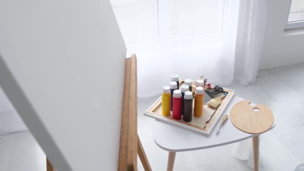 Pots avec des peintures colorées os et spatules se tiennent sur le bureau des artistes près du chevalet avec toile blanche. Contenants en plastique avec différentes peintures appartenant à l'artiste. L'atelier est très lumineux. - Séquence, vidéo