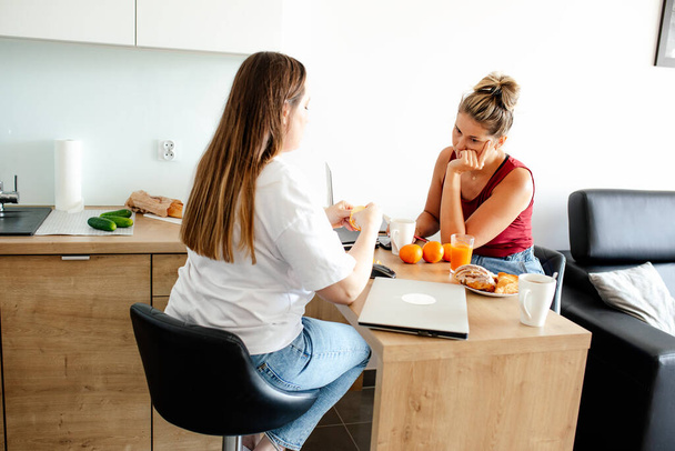 Communication and mutual understanding. Two cute girlfriends are sitting facing each other in kitchen talking, having lunch with fruit, laptop is lying next to them. Comfort and work. Home office,. - Photo, image
