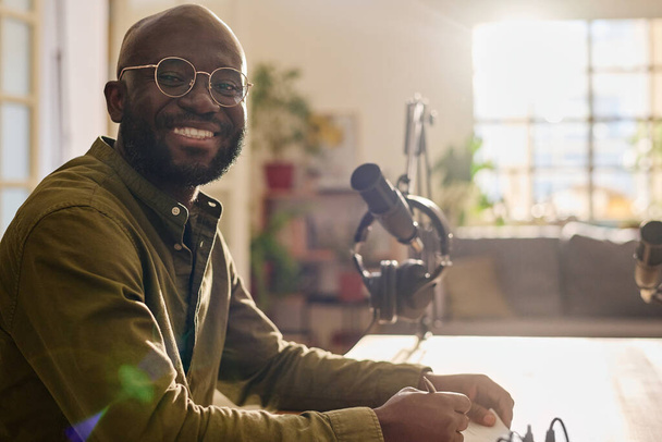 Mladý veselý Afroameričan v brýlích dívá na kameru, zatímco sedí na pracovišti a zapisuje plán diskuse - Fotografie, Obrázek