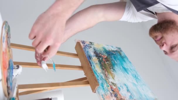Вертикальное видео профессионального художника, рисующего картину на белом холсте с помощью кистей и мольберта. Процесс создания картины художником - Кадры, видео