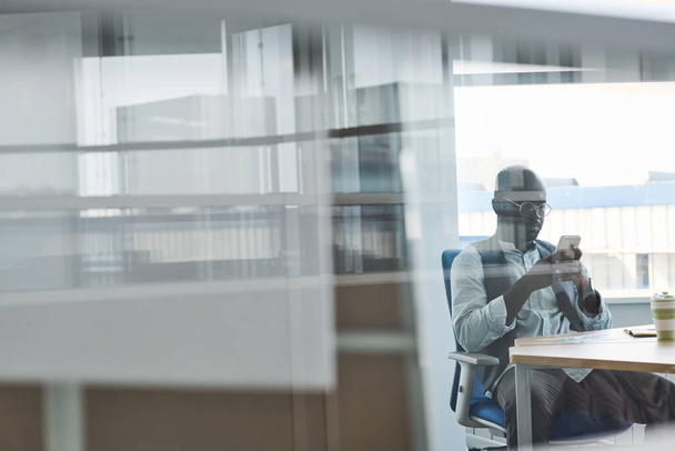 Молодой афроамериканский бизнесмен в формальной одежде, прокручивающийся в смартфоне, сидя на рабочем месте в офисе с прозрачными стенами - Фото, изображение
