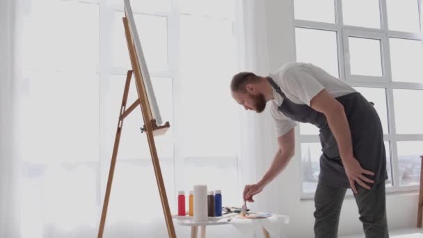 Een getalenteerde mannelijke kunstenaar, met behulp van een borstel, creëert een modern meesterwerk van olieverf op een wit doek. Het doek ligt op een ezel in een grote studio. - Video