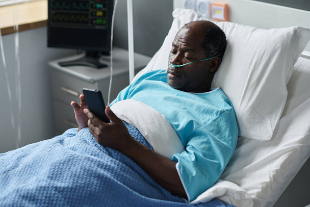 病院病棟のベッドに横になっている間にスマートフォンにメッセージを入力する高齢者 - 写真・画像