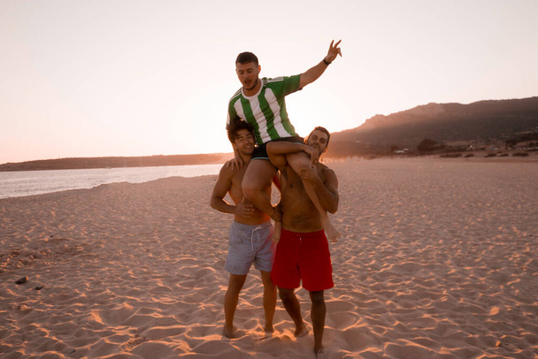 Dwoje młodych przyjaciół hiszpańskiego pochodzenia podnosi swojego przyjaciela ramionami, ciesząc się wakacjami na zacisznej plaży nad morzem i zachodem słońca.. - Zdjęcie, obraz