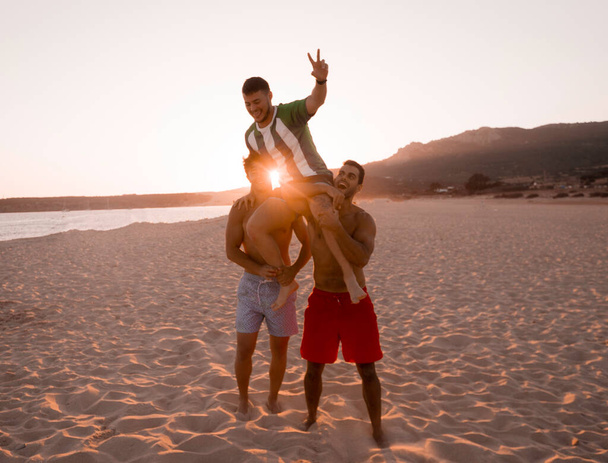 A két fiatal barna barát felemeli a barátját a karjával, miközben élvezik a nyaralást egy félreeső strandon.. - Fotó, kép