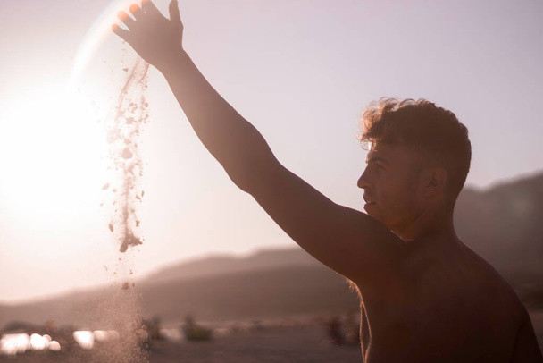 Egy fiatalember gyengéden leejti a homokot az ujjai között, felemeli a karját a nyaralás napján a parton, háttérvilágítás van rajta.. - Fotó, kép