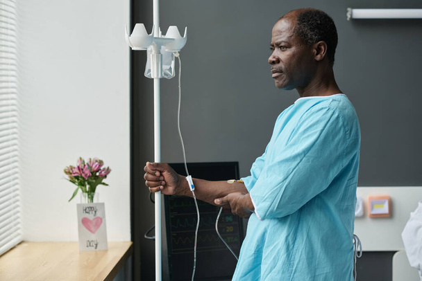 Paciente anciano afroamericano de pie con gotero en la sala y mirando a través de la ventana con expresión pensativa - Foto, Imagen