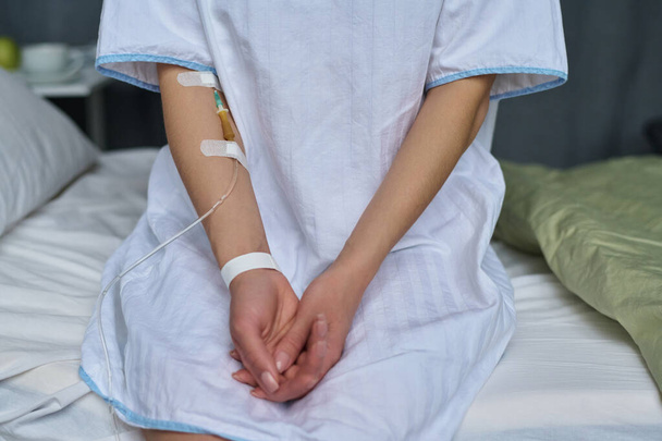 Close-up of young patient with catheter in her arm sitting on bed in ward - Foto, afbeelding