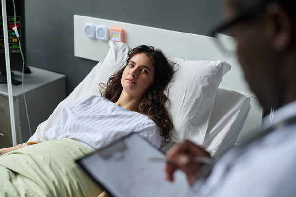 Young woman talking to doctor about her health condition while lying on bed in ward - Photo, Image