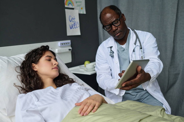 Афроамериканский врач показывает диагноз на экране на цифровом планшете пациентке, пока она лежит на кровати в палате - Фото, изображение