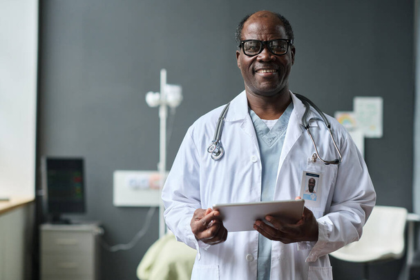 Porträt eines afroamerikanischen Arztes im weißen Mantel, der in die Kamera lächelt, während er auf der Station ein digitales Tablet benutzt - Foto, Bild