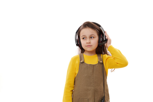 Sevimli bir ilkokul öğrencisi, 5-6 yaşlarında, günlük giysiler içinde, ses kulaklığı takan, izole beyaz bir geçmişi olan sevimli bir kız çocuğu. Çevrimiçi iletişim. Uzaktan eğitim. E-öğrenme - Fotoğraf, Görsel