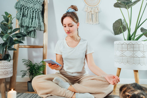 Young healthy woman using phone for setting mobile app for meditation at home, sitting in lotus position on floor cushion with relaxed cat pet in modern Scandi interior home. Meditating, relaxed. - Foto, Imagem