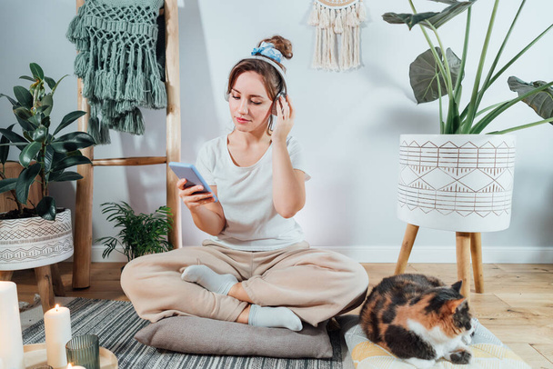 Young healthy woman using phone for setting mobile app and headset for meditation at home, sitting in lotus position on floor with relaxed cat pet in modern Scandi interior home. Meditating, relaxed - Foto, afbeelding