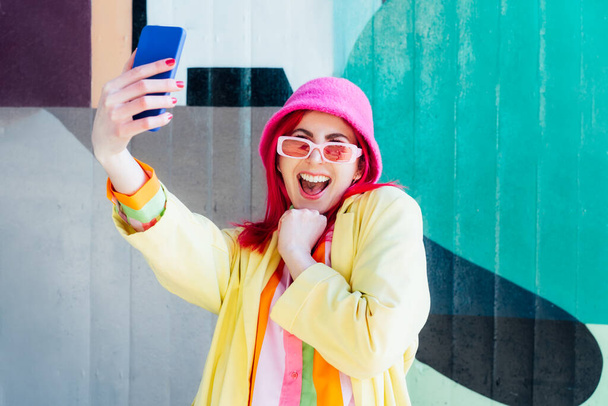 Excited pink hair woman screaming while taking a selfie photo outdoors. Emotional hipster fashion woman in bright clothes, pearl pink sunglasses, bucket hat taking selfie photo on the phone camera. - Photo, image