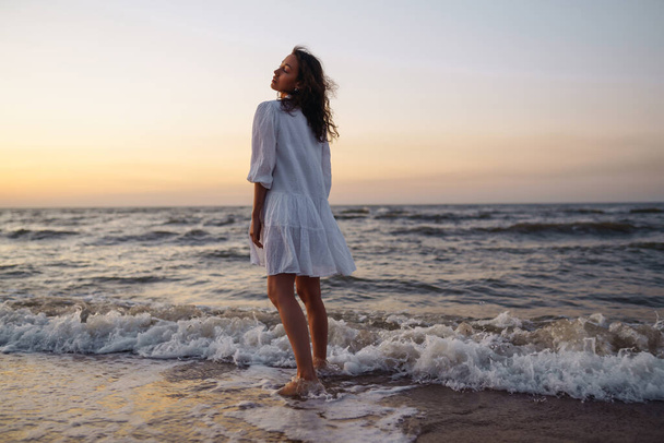 Beautiful model on the seashore posing in a delicate white dress. A young woman enjoys an incredible sunrise on the beach. Lifestyle. Vacation, travel concept.  - Photo, image