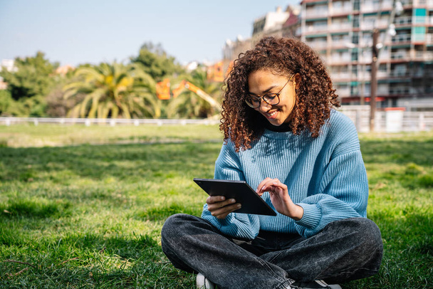 Cheerful young curly haired Hispanic female in casual wear and eyeglasses sitting on grassy lawn in wireless headphones while browsing tablet with toothy smile in sunny park - Fotó, kép