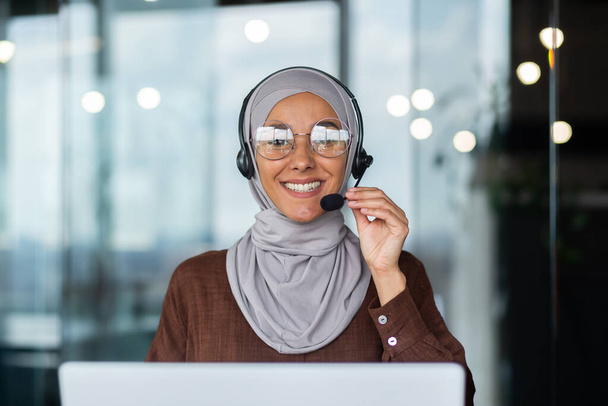 Close-up photo. Young hispanic woman in hijab sitting in office in headset and talking, consulting, advising on video call from laptop. He holds the microphone in his hand, looks at the camera. - Foto, Imagem