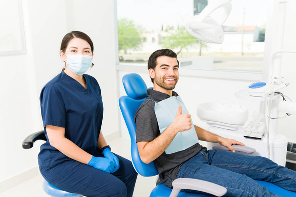 Задоволений щасливий пацієнт чоловічої статі робить великі пальці вгору, відчуваючи себе щасливим з його стоматологічним лікуванням після приходу до стоматолога
 - Фото, зображення