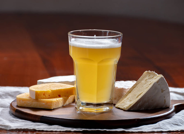 Glass of Belgian white unfiltered beer with notes of coriander and orange peer and wooden board with variety of belgian cheeses, beer and cheese pairing - Photo, Image