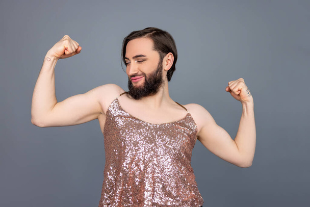 Satisfied proud transsexual person wearing shiny tank top showing biceps and his power expressing pride posing isolated over gray background - Фото, изображение