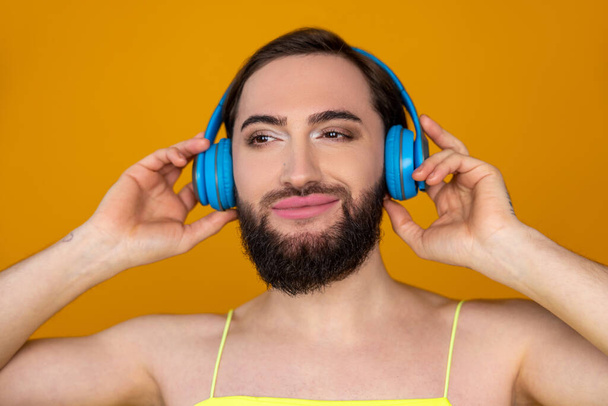 Closeup portrait of smiling satisfied transgender person with beard listening to music via blue headphones enjoying posing isolated over orange background - Foto, imagen