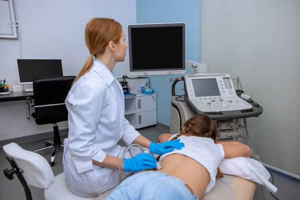Female doctor performing kidney ultrasound scan on school aged girl medical specialist examining child back with sonogram equipment in hospital. - Φωτογραφία, εικόνα