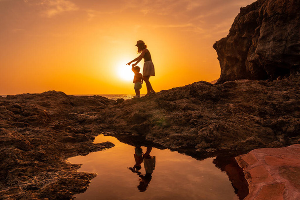 Silhouette of mother and son walking in the sunset on the beach of Tacoron in El Hierro, Canary Islands, orange sunset, walking along the sea pointing the path - Photo, image