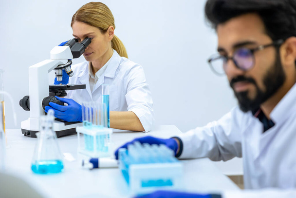 Team van wetenschappers onderzoekt monster, vrouwelijke arts met behulp van microscoop, mannelijke onderzoeker werkt met chemische vloeistoffen in kolf in modern lab. - Foto, afbeelding