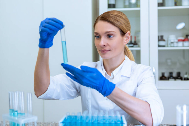 Blonde successful woman researcher working with medical samples, holding test tube with blue liquid, making microbiology investigating in laboratory. - Fotó, kép