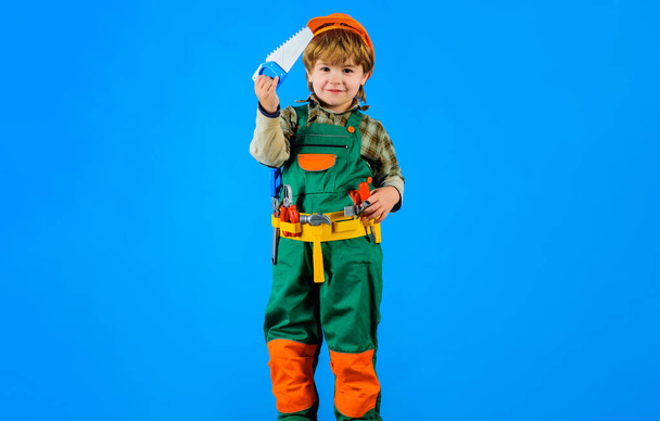 Construtor menino em capacete de segurança e cinto de ferramentas com serra. Criança em uniforme construtor com cinto de ferramentas. Ferramentas para construção. Reparador de crianças com serra. Trabalhar com ferramentas de reparo. Pequeno rapaz trabalhador da construção - Foto, Imagem