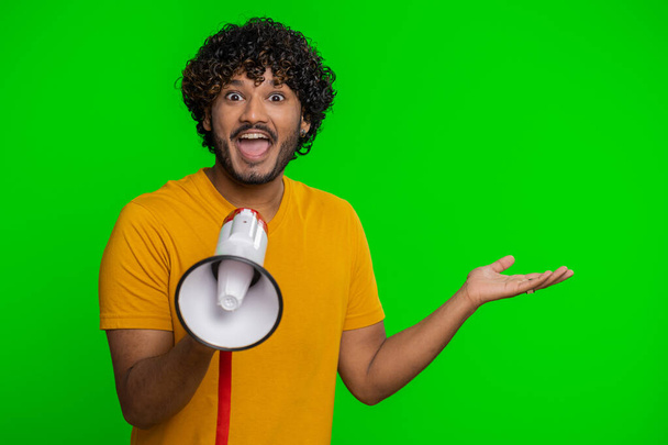 Indian man talking with megaphone, proclaiming news, loudly announcing advertisement, warning using loudspeaker to shout speech, pointing empty place. Copy-space. Handsome guy on chroma key background - Foto, Imagen